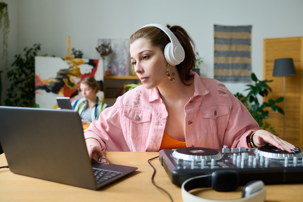 A young woman in a pink denim jacket with a ponytail on her head and white headphones sitting at a table in a bright room uses a laptop and a music console creating tracks